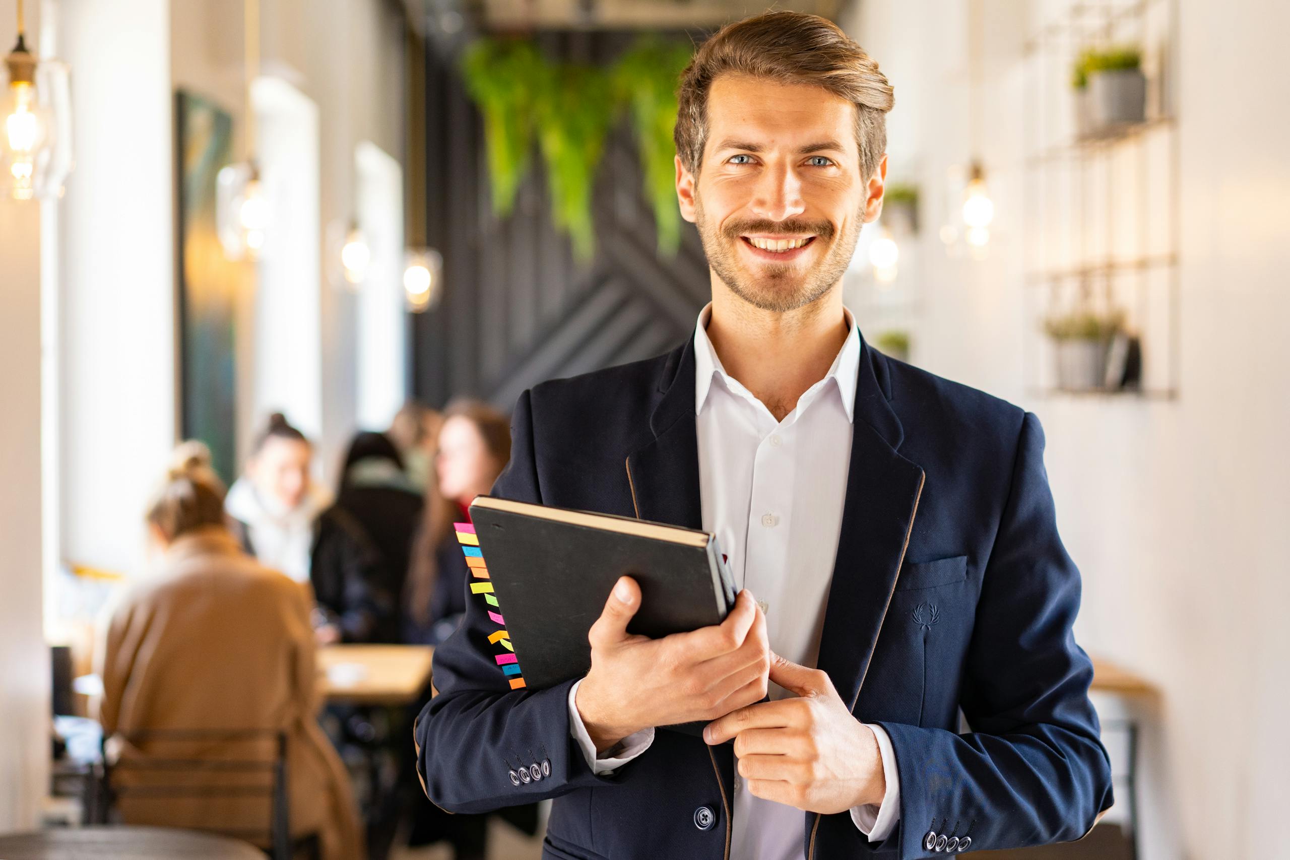 Smiling businessman in a blue suit holding a notebook in a modern office setting.